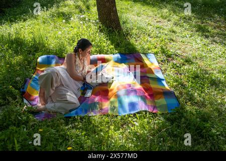 Una bella donna di mezza età riposa e legge un libro, distesa in mezzo a un campo su una coperta colorata a scacchi Foto Stock