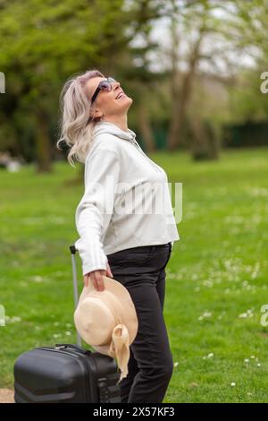 Donna con una valigia sorridente, in piedi in un parco. Foto Stock