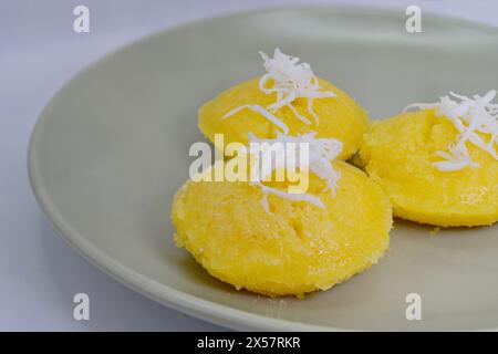 Tre Khanom Tan di colore giallo brillante o Thai Toddy Palm Cakes, con condimenti di cocco grattugiato, su piatto da portata verde Foto Stock