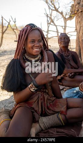 La giovane donna Himba, vestita tradizionalmente, seduta all'ombra, vicino a Opuwo, Kaokoveld, Kunene, Namibia Foto Stock