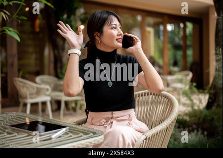 Una donna asiatica bella e positiva siede ad un tavolo all'aperto di un bar che parla al telefono mentre saluta la mano per chiamare o salutare qualcuno. persone e l Foto Stock