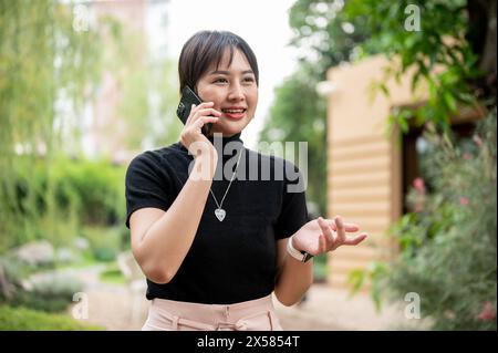 Una donna asiatica attraente e positiva in abiti casual sta parlando al telefono con qualcuno mentre cammina in un bellissimo giardino. Foto Stock