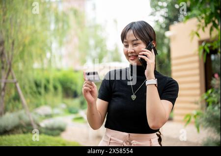 Una bella donna asiatica in abiti casual chiama il call center della sua carta di credito o di debito mentre cammina in giardino. Foto Stock