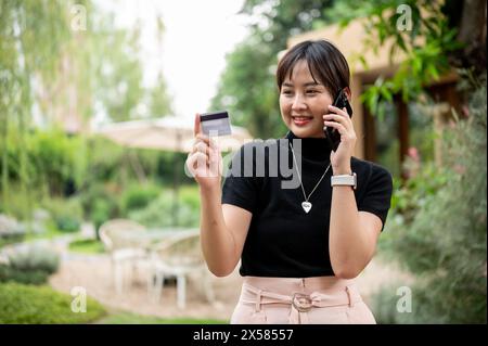 Una bella donna asiatica in abiti casual chiama il call center della sua carta di credito o di debito mentre cammina in giardino. Foto Stock