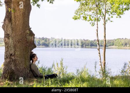 Un'immagine di riposo tranquillo, che mostra una donna seduta sotto il robusto tronco di un albero. Il lago sullo sfondo, incorniciato da una vegetazione verdeggiante, accentua la sensazione di solitudine e il legame con la natura. Momento tranquillo vicino al lago: Donna che riposa sotto un albero. Foto di alta qualità Foto Stock