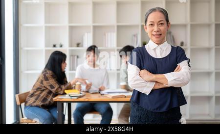 Un ritratto di una professoressa asiatica anziana sorridente e soddisfatta in aula con le braccia incrociate mentre gli studenti studiano. lecturi Foto Stock