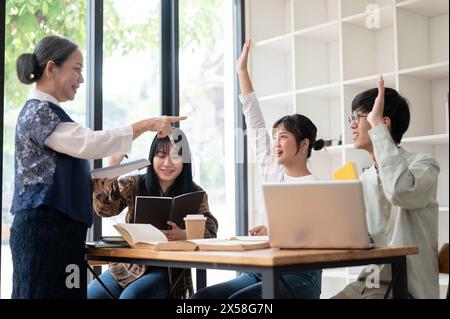 Due giovani studenti universitari asiatici stanno alzando la mano per rispondere a una domanda dell'insegnante, studiando in classe con un matur esperto Foto Stock