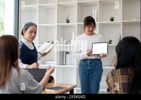 Una giovane studentessa asiatica sta mostrando il suo progetto sul suo tablet digitale, presentando il suo progetto in classe al suo insegnante e agli amici. Foto Stock