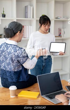 Una giovane studentessa asiatica sta mostrando il suo progetto sul suo tablet digitale, presentando il suo progetto in classe al suo insegnante e agli amici. Foto Stock