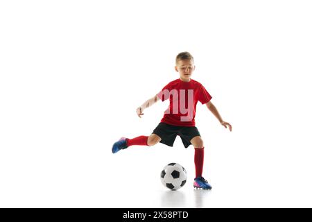 Il colpo dinamico di un bambino in uniforme sportiva gioca a calcio, allenando la palla passante su sfondo bianco. Foto Stock