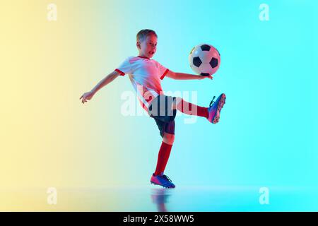 Il tiro dinamico di un bambino in uniforme sportiva gioca a calcio, allenandosi passando la palla in luce al neon su sfondo gradiente. Foto Stock