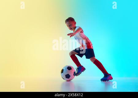 Atletico e sportivo ragazzo in uniforme da allenamento tecnica dribbling in movimento con luce al neon su sfondo gradiente. Scatto dinamico. Foto Stock