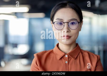 Un ritratto professionale di una giovane donna d'affari asiatica che indossa occhiali e una camicetta arancione, in piedi con sicurezza in un ambiente d'ufficio contemporaneo. Foto Stock