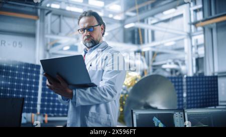 Ingegnere maschile pensa con fiducia e concentrazione, lavorando su notebook presso lo stabilimento di produzione di satelliti aerospaziali. Top World Scientists doing Science and Technology Research in Space Program Foto Stock