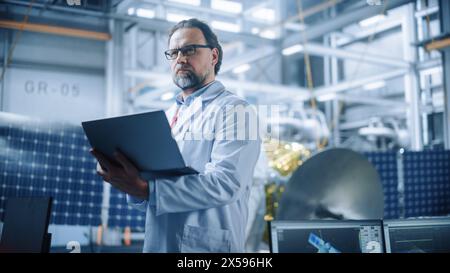 Ingegnere maschile pensa con fiducia e concentrazione, lavorando su notebook presso lo stabilimento di produzione di satelliti aerospaziali. Top World Scientists doing Science and Technology Research in Space Program Foto Stock