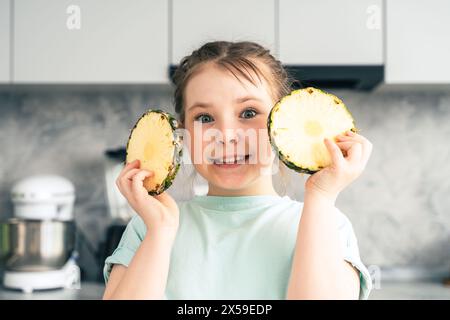 Allegra bambina con ananas tagliato in cerchio in cucina a casa. Guarda nella cornice e sorride. Messa a fuoco selezionata. Alta qualità Foto Stock