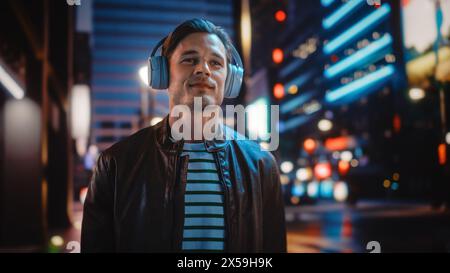 Ritratto dell'uomo bello che indossa le cuffie passeggiando per Night City Street piena di luce al neon. Uomo sorridente ed elegante che ascolta musica, podcast, talk show. Foto Stock