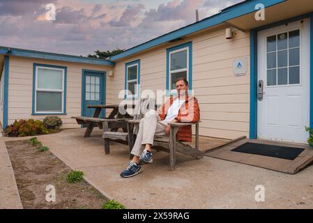 Persona in giacca arancione che si rilassa sulla panca di legno presso il Blue trimmed Building nella costa della California Foto Stock