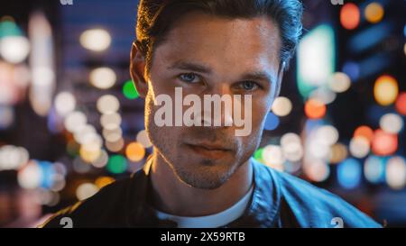 Ritratto dell'uomo bello e serio che guarda la macchina fotografica, in piedi nella città notturna con bokeh Neon Street Lights sullo sfondo. Giovane e sicuro di sé con capelli eleganti. Primo piano Foto Stock