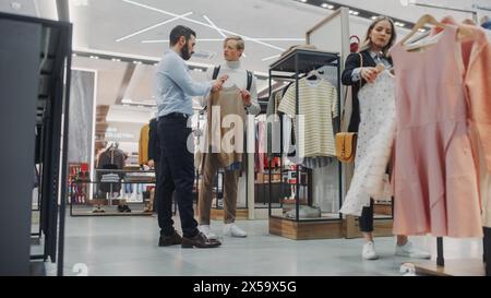 Bel cliente che fa acquisti nel negozio di abbigliamento, Retail Sales Assistant aiuta con consigli. Persone diverse nel negozio alla moda, scelta di abiti eleganti, marchio colorato con design sostenibili Foto Stock