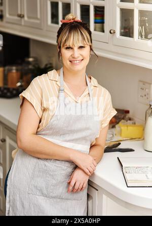 Sorridi, donna e cuoci in cucina per ritratti con ricettario, ingredienti e attrezzature con grembiule per dessert. Felice, chef donna e lettura per Foto Stock