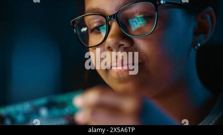 Primo piano Ritratto di una giovane ragazza nera multietnica adolescente carina sta studiando un piccolo oggetto per circuito stampato. La giovane donna indossa occhiali e lavora al suo School Science Project. Foto Stock