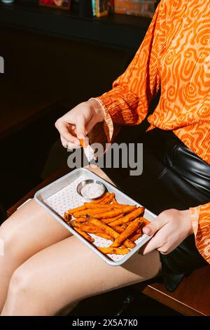 Una donna corta e irriconoscibile sta gustando spicchi di patate dolci fritte servite con una cremosa immersione al tartufo in un ambiente informale. Foto Stock