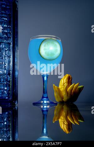 Un vivace cocktail blu con una fetta di lime, accompagnato da agrumi di buddha, su una superficie riflettente Foto Stock