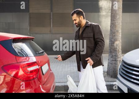 Un maschio adulto sblocca la sua auto rossa con una chiave elettronica dopo essere uscito da un supermercato, portando con sé sacchetti di plastica bianchi Foto Stock