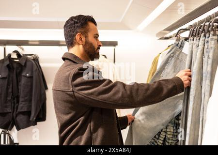 Un uomo in giacca casual acquista jeans in denim in un negozio di abbigliamento ben illuminato, esaminando attentamente le sue opzioni Foto Stock