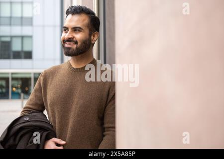 Un uomo dall'abbigliamento casual sorride mentre si trova in città, con un cappotto e un look ottimista Foto Stock