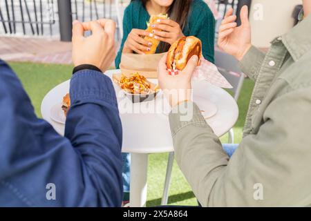 Un gruppo di amici è riunito intorno a un tavolo, gustando hamburger e patatine fritte da un camion del cibo, che rappresenta un pasto urbano informale Foto Stock