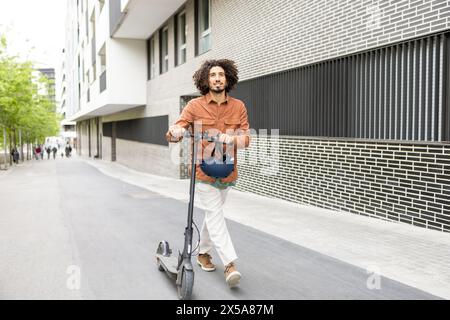 Un giovane uomo elegante con capelli ricci guida uno scooter elettrico su una strada cittadina, intrecciando un ambiente urbano contemporaneo con edifici moderni Foto Stock
