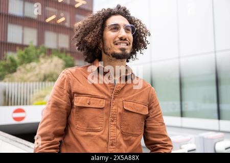 Un giovane uomo con capelli ricci e occhiali sguardi attentamente, vestito con un'elegante camicia marrone su uno sfondo urbano Foto Stock