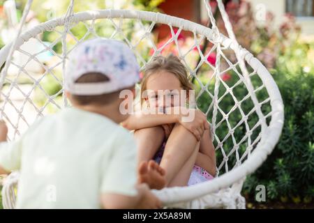 Due giovani fratelli si stanno impegnando in un gioco allegro su un'altalena bianca in un giardino verde, crogiolandosi nel calore di una giornata di sole e condividendo un momento di happ Foto Stock