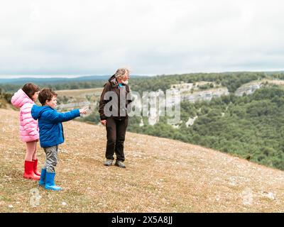 Nonno con nipoti, un ragazzo e una ragazza, esplorando la natura insieme su una collina, puntando su una vista panoramica. Foto Stock