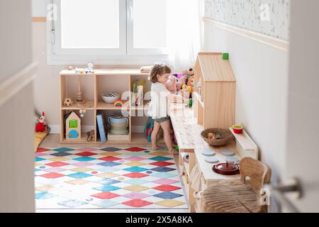 Una giovane ragazza ama divertirsi all'interno di una vivace sala giochi illuminata dal sole piena di giocattoli e di un tappeto geometrico colorato Foto Stock