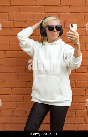 Una donna bionda in un abbigliamento casual usa lo smartphone indossando le cuffie contro una parete urbana in mattoni. Foto Stock