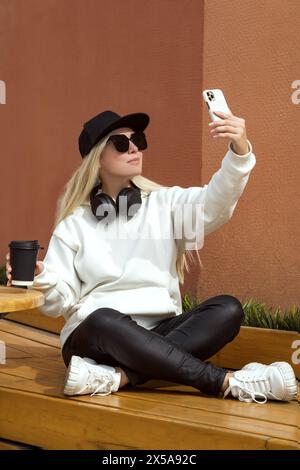 Donna bionda alla moda in stile casual, seduta su una panchina e scattata un selfie con un telefono cellulare in un ambiente urbano. Foto Stock
