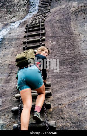 Da sotto una donna con uno zaino sale una pericolosa scala a catena su una montagna rocciosa, mostrando l'avventura e la sfida dell'escursionismo. Foto Stock