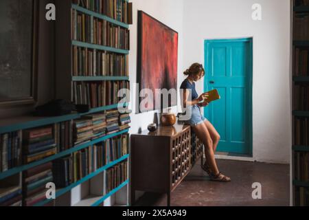 Una giovane donna si appoggia contro una credenza, infilata in un libro accanto a uno scaffale pieno di libri, vicino a una porta di tè. Foto Stock