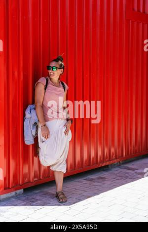 Una donna allegra con un abbigliamento casual estivo, occhiali da sole e sandali si appoggia su una vivace parete rossa, guardando lontano dalla fotocamera con un espresso gioioso Foto Stock