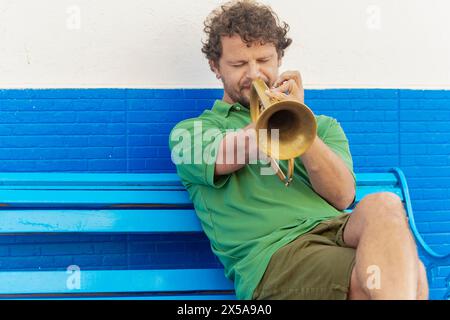 Un uomo con un braccio suona con passione la tromba seduto su una panchina blu, gli occhi chiusi, perduti nella musica. Foto Stock