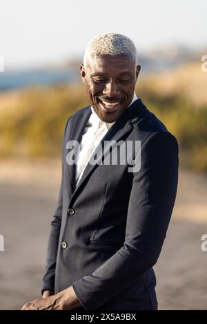 Un elegante maschio afroamericano con un affascinante sorriso, vestito con un costume classico, si mette in posa rilassata sullo sfondo di una spiaggia sabbiosa Foto Stock