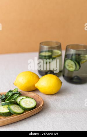 Due bicchieri riempiti di cetriolo e acqua infuso al limone, esposti con limoni freschi e fette di cetriolo su un vassoio di legno, promuovendo un saluto sano Foto Stock