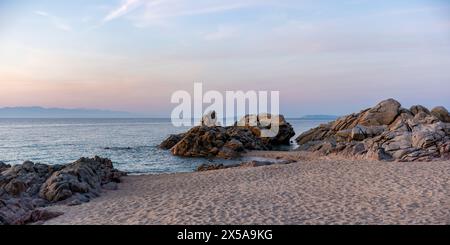 Un calmo tramonto proietta sfumature morbide sull'idilliaca spiaggia di sabbia e le scogliere aspre di una tranquilla baia in Sardegna, Italia, con un mare sereno e un cielo limpido Foto Stock