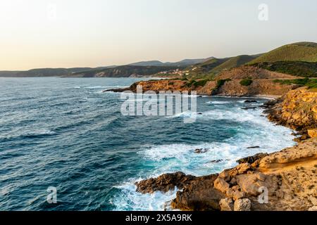 La luce del sole dorata bagna la costa frastagliata della Sardegna, in Italia, con onde che si infrangono contro le coste rocciose, mettendo in evidenza la bellezza selvaggia del Mediterraneo Foto Stock
