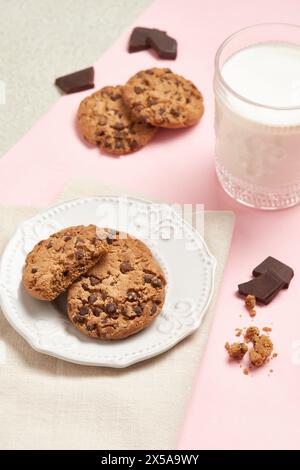 Vista dall'alto di deliziosi biscotti con scaglie di cioccolato su un piatto bianco con un bicchiere di latte e pezzi di cioccolato sparsi su sfondo rosa Foto Stock
