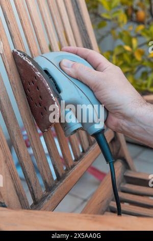 Primo piano della mano di un uomo con una levigatrice elettrica per levigare e rifinire la superficie di una sedia di legno, preparandola per la verniciatura Foto Stock