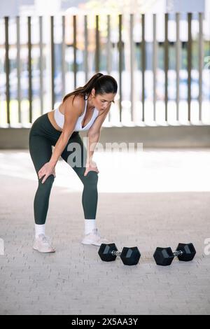 La donna in forma si mette in pausa, si mette le mani sulle ginocchia, dopo essersi allenata con i manubri a casa, mostrando uno stile di vita attivo e una routine di fitness indoor Foto Stock